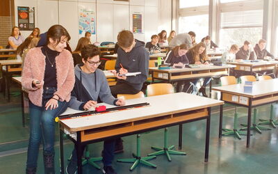 SchülerInnen in einem Klassenzimmer, umgeben von Tischen, Stühlen und anderen SchülerInnen, arbeiten zusammen an ihrem Versuch.