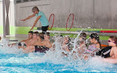 Eine Gruppe von SchülerInnen übt im Pool, während die Lehrerin ihnen die Beinarbeit fürs Schwimmen erläutert.