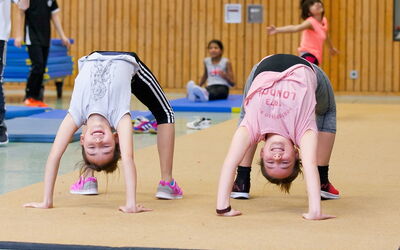 Zwei junge Mädchen machen eine Rückenbrücke in der Sporthalle und lachen dabei.