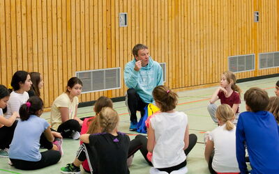 Sportlehrer unterrichtet eine Gruppe von SchülerInnen, die auf dem Boden sitzen.