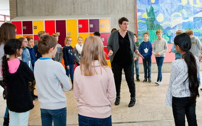 SchülerInnen versammeln sich um eine Lehrerin in einem Klassenzimmer, während sie gespannt zuhören.