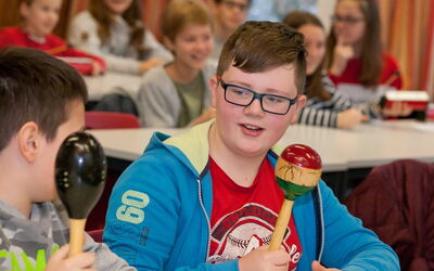 Schüler mit Brille und rotem Shirt spielt begeistert Rasseln im Klassenzimmer.